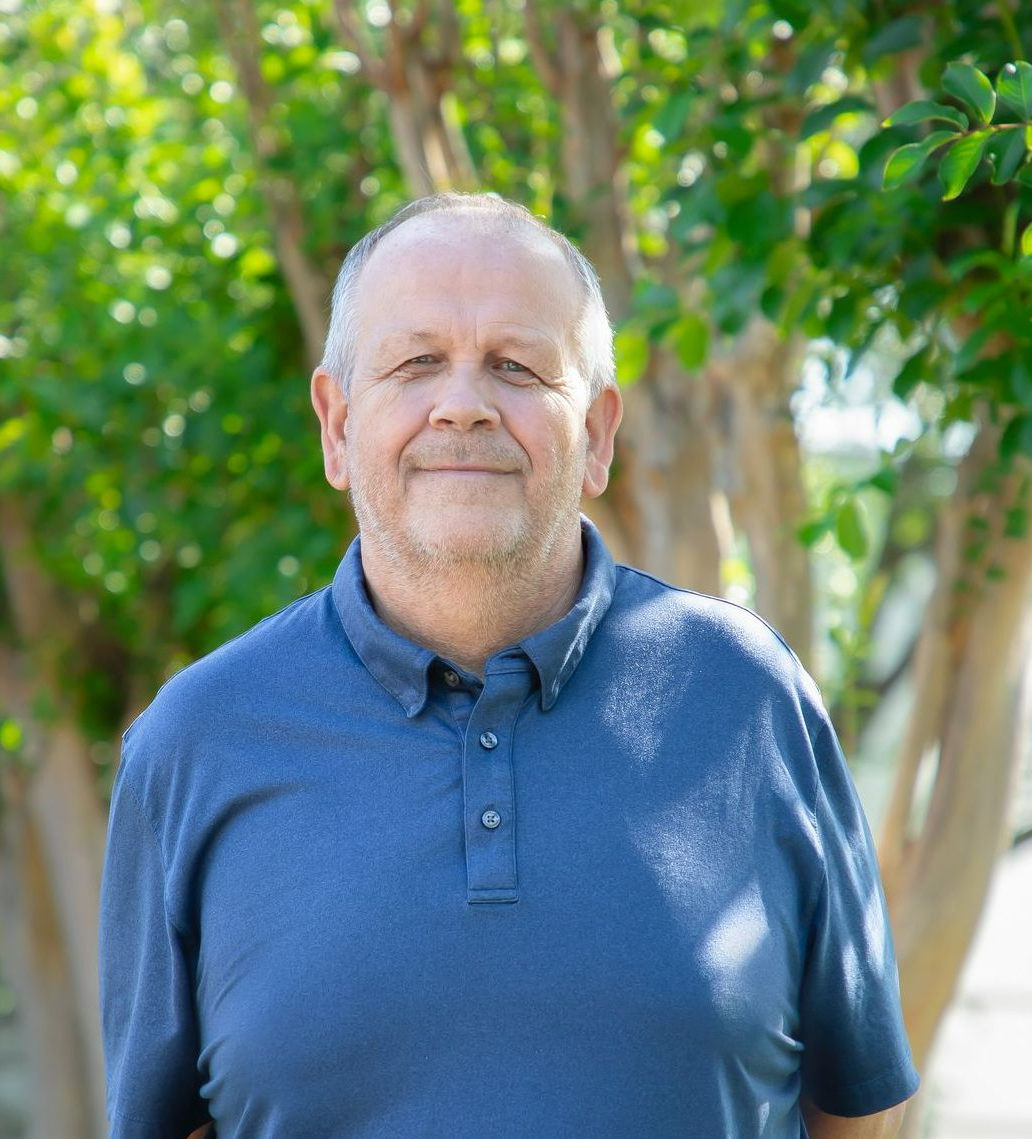 Man in blue polo smiling outdoors, trees in background.