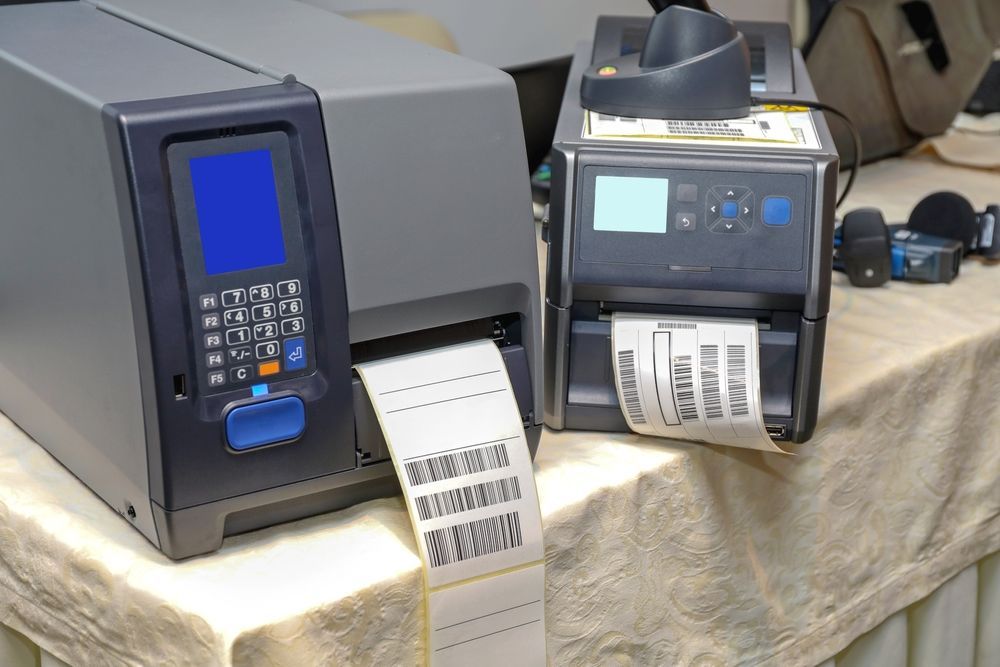 Two label printers on a table printing barcodes. One is gray, the other is black.