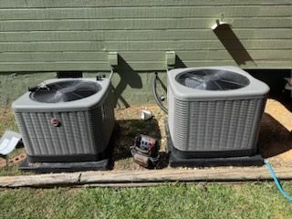 Two grey HVAC units sit on black pads against a green wall with a tool bag between them.