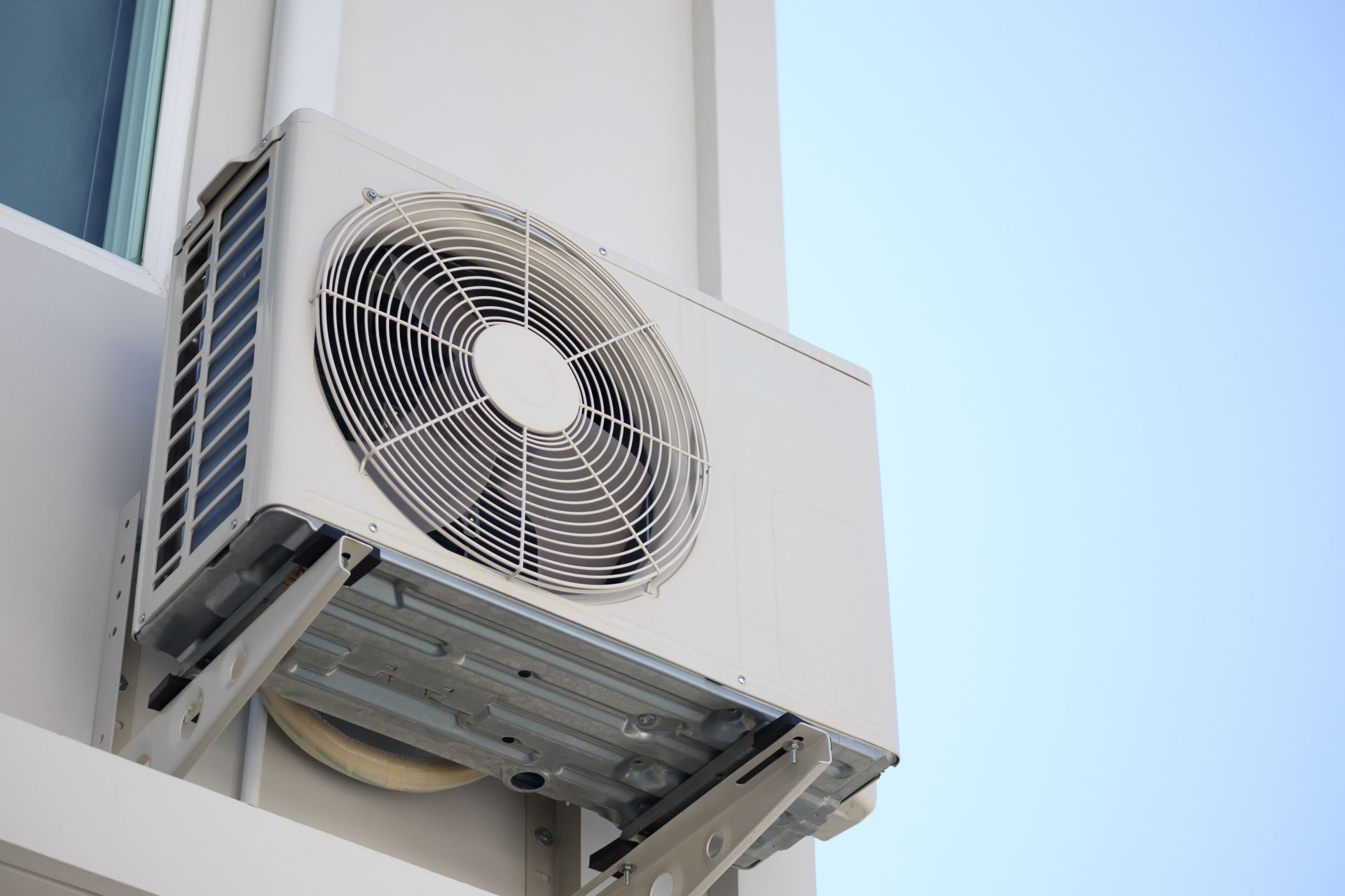 A white air conditioning unit mounted on the exterior wall of a building against a light blue sky.