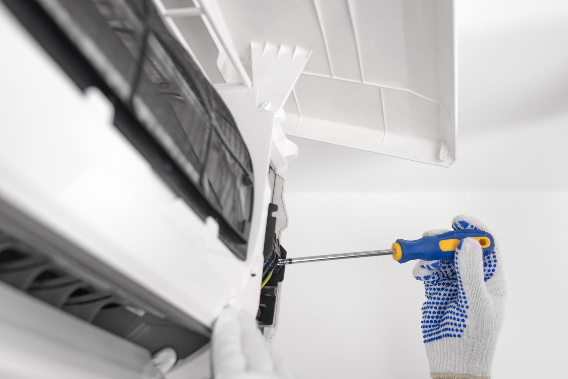 A person wearing work gloves uses a screwdriver to service the interior components of a wall-mounted air conditioner.