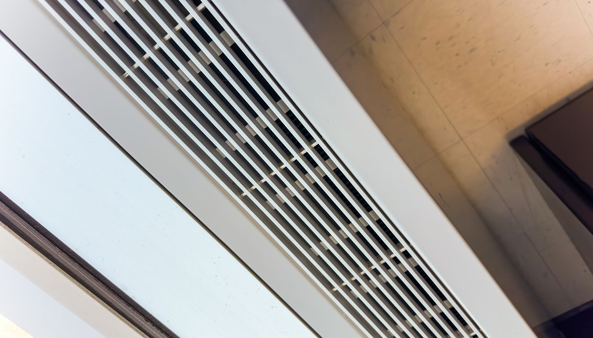 A white, slatted linear HVAC air vent mounted on a ceiling near a window.