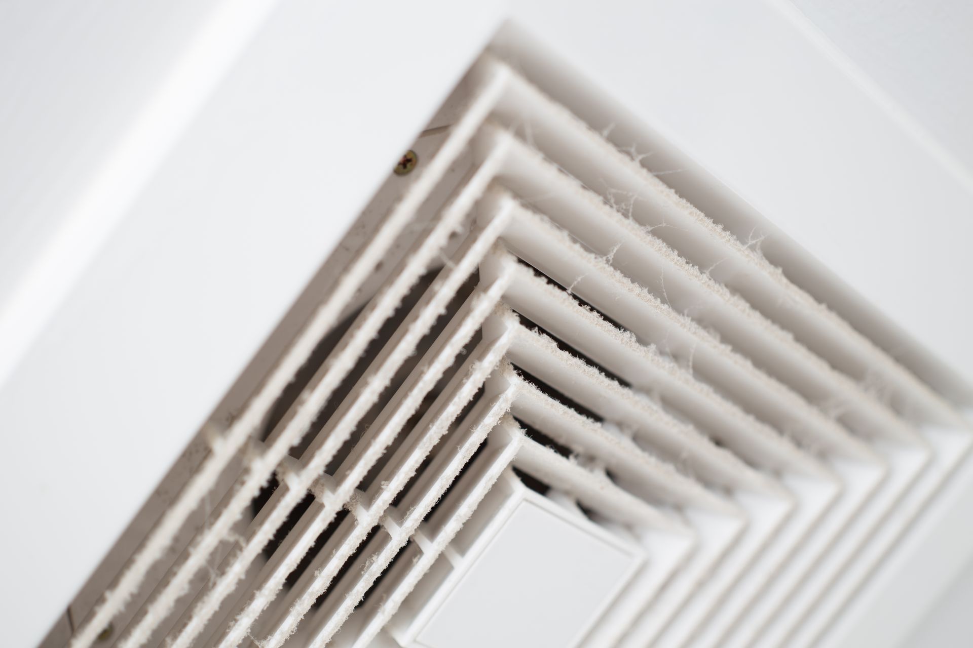 A close-up, angled view of a white, dusty ceiling air vent with multiple slatted layers.