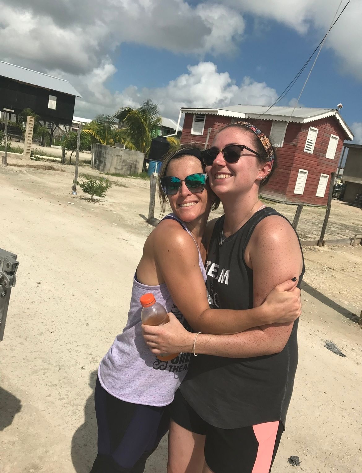 Two women hugging outdoors, smiling. One holds a drink. Buildings and sky in the background.