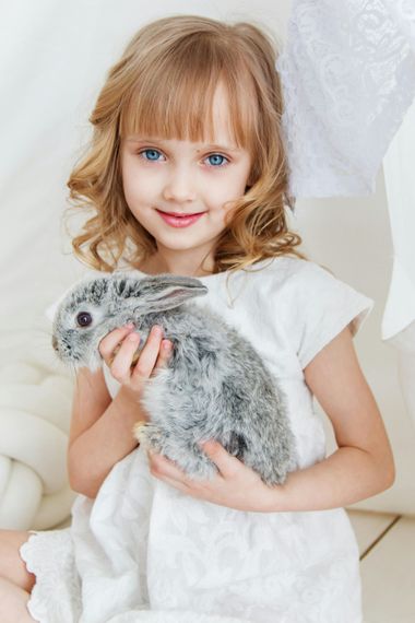 Girl with blue eyes, holding a gray rabbit, smiling. She is wearing a white dress.