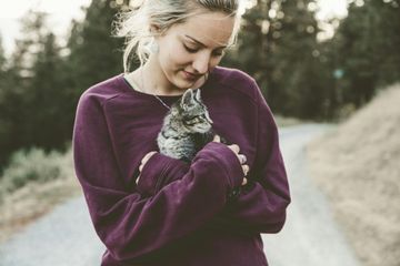 Woman in purple sweatshirt holding a small gray cat on a path in a wooded area.