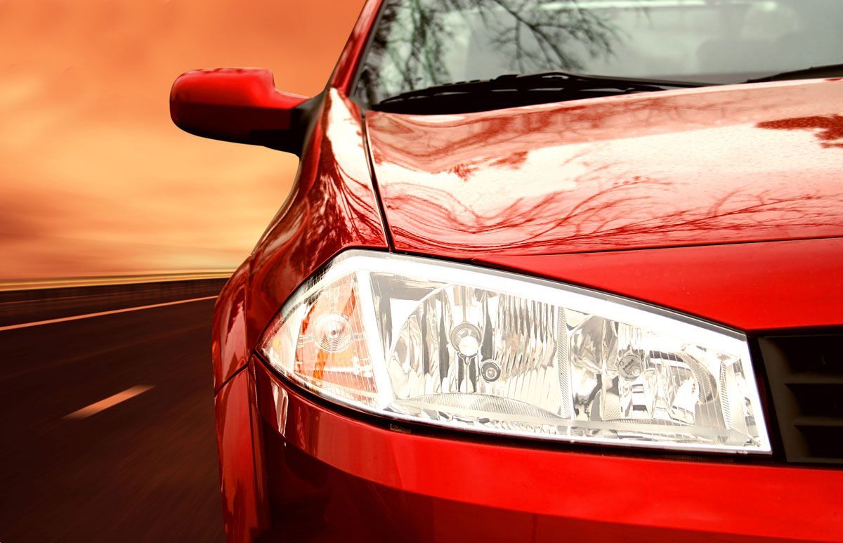 Red car driving on road, headlight lit, against orange sky.