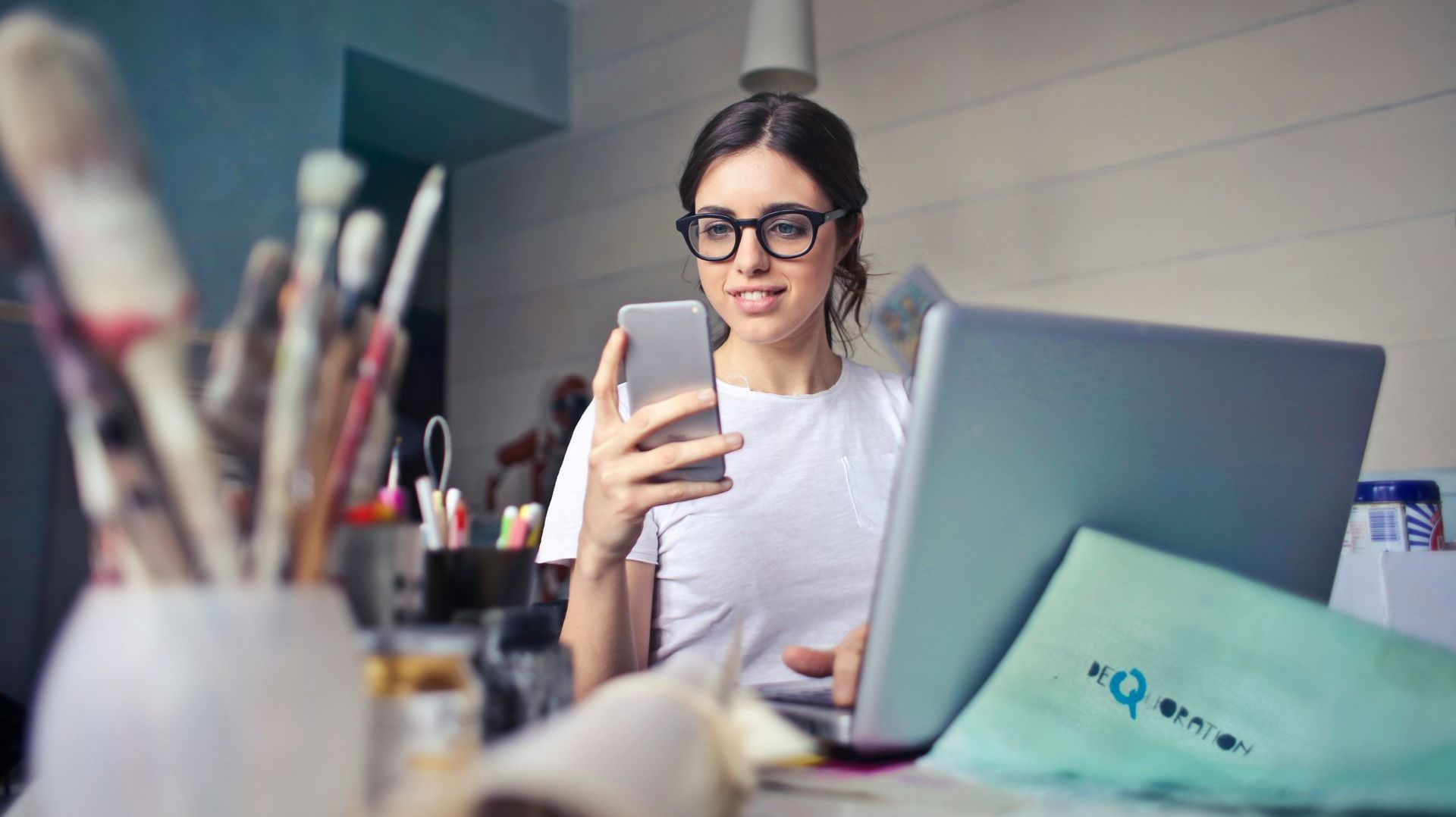 A woman is sitting at a desk using a laptop and a cell phone.