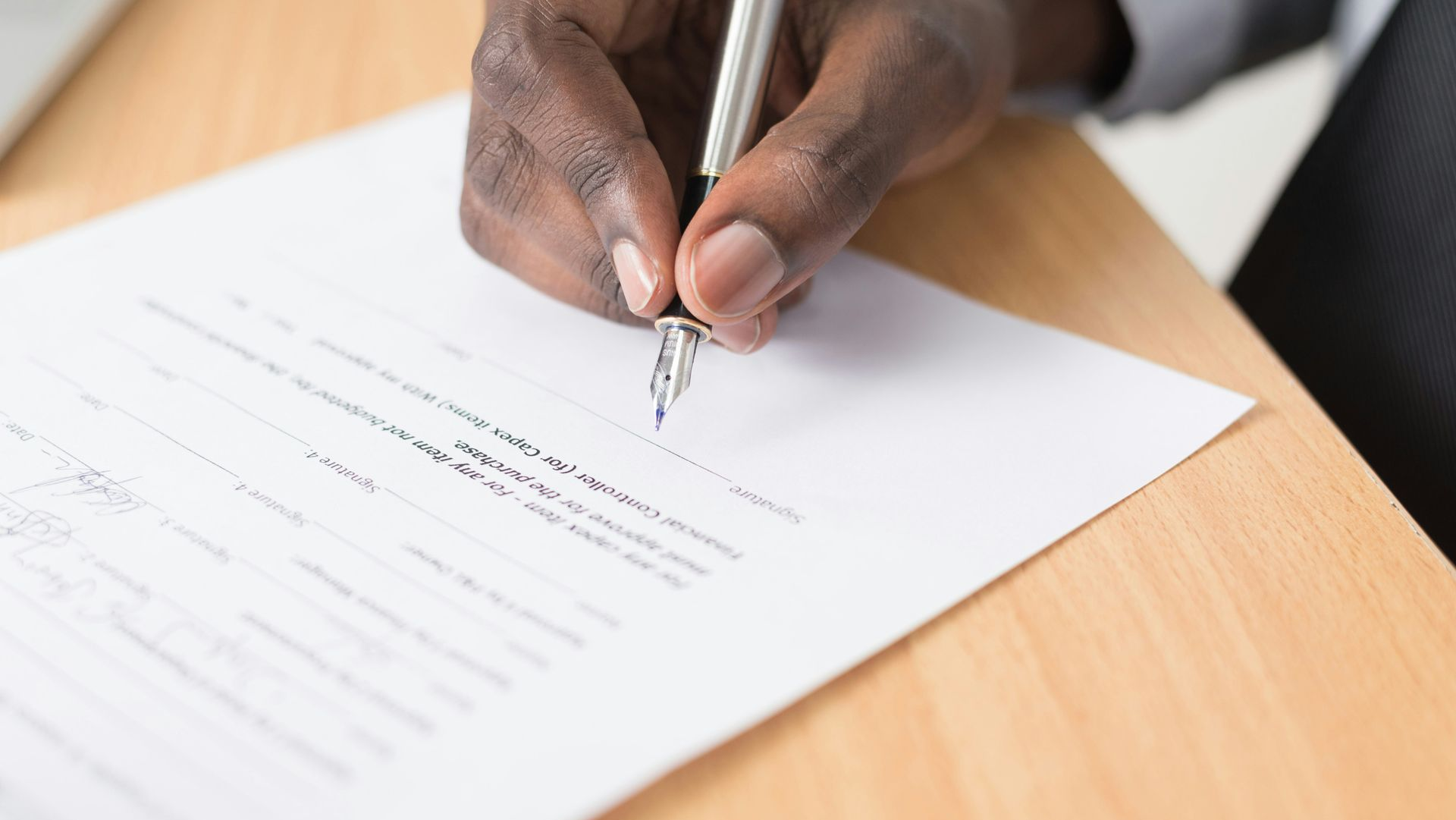 Person's hand signing a document with a pen on a wooden surface.