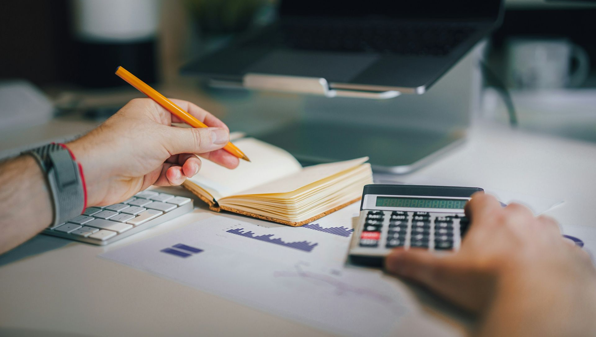 Person using a calculator and writing in a notebook next to a laptop on a stand.