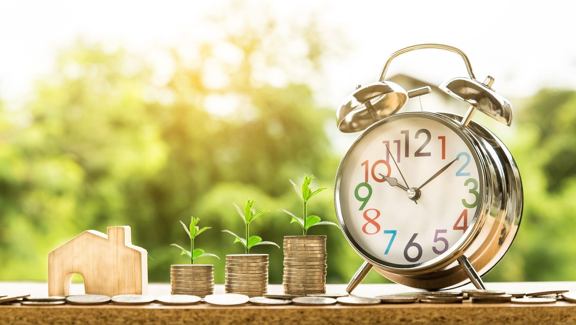 An alarm clock is sitting on a table next to stacks of coins and a house.