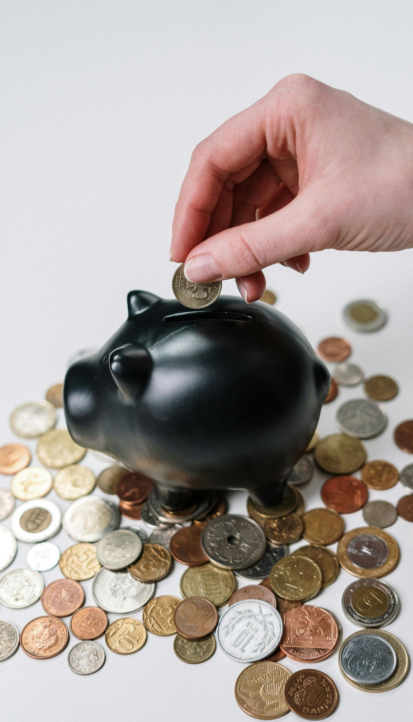 Hand placing a coin into a black piggy bank surrounded by various coins on a white surface.