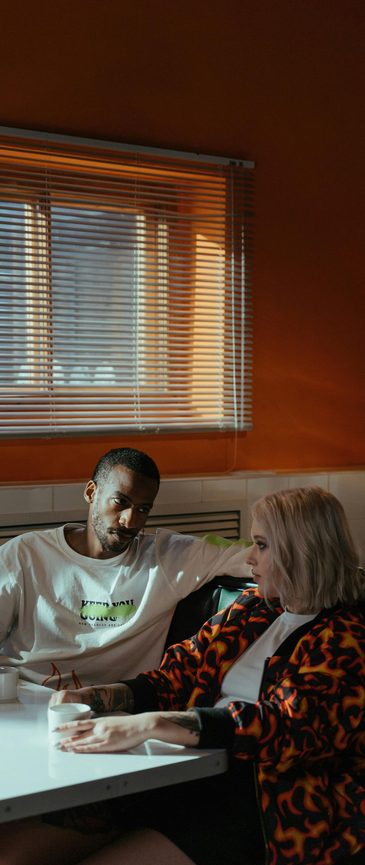 Two people sitting at a table in an orange room. The woman wears a patterned jacket, the man a white shirt.