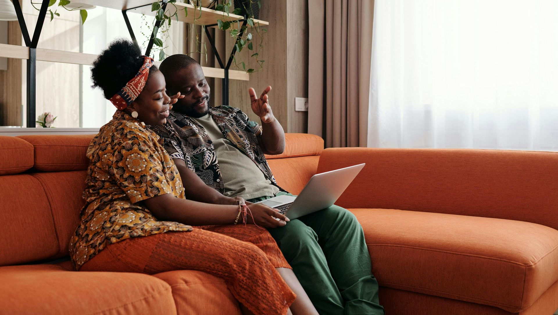 Couple on orange couch looking at laptop, smiling. Green pants, floral top, wooden shelves with plants in a room.