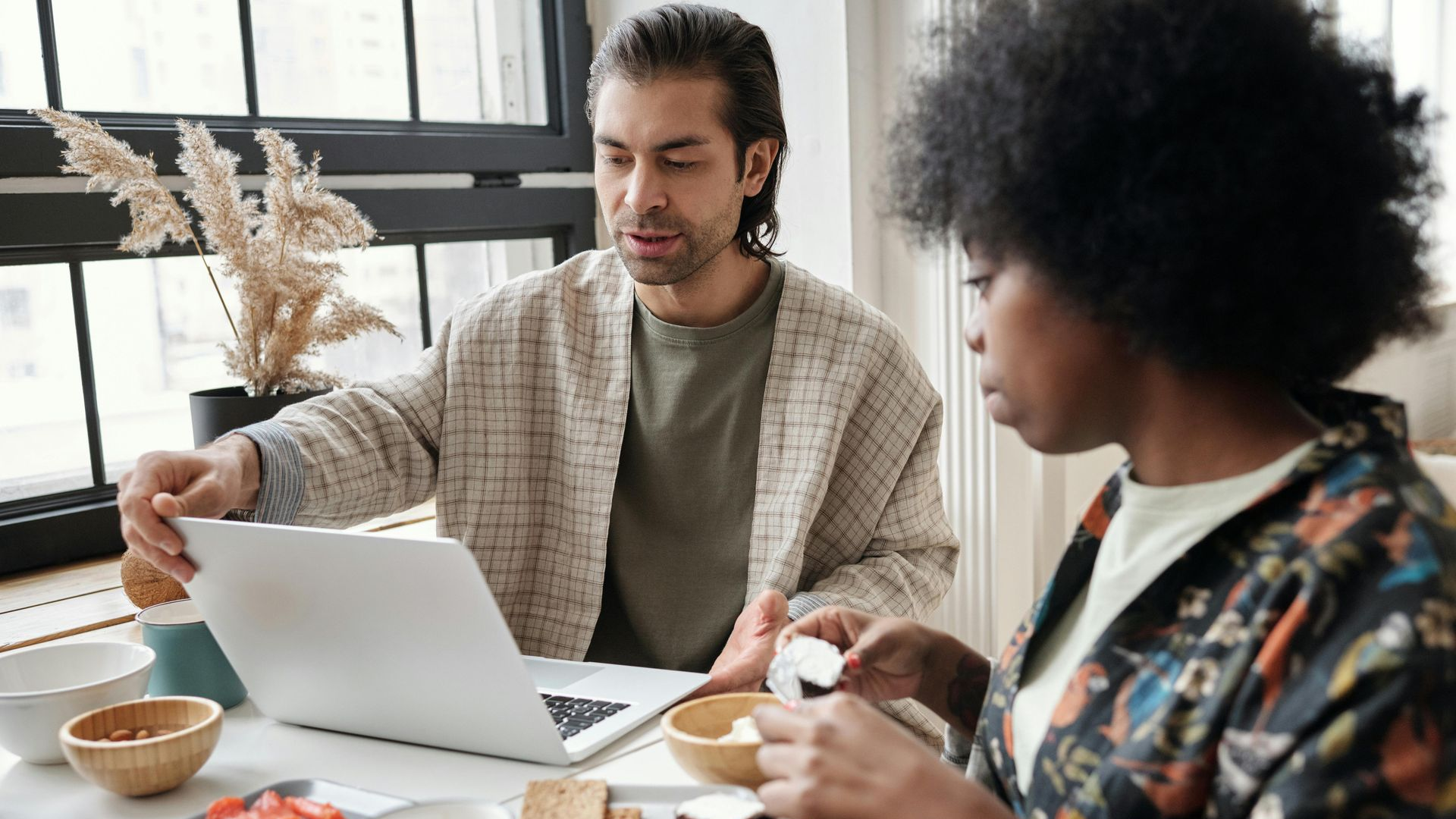 A man and a woman at a table with a laptop and breakfast items. The man points at the screen.