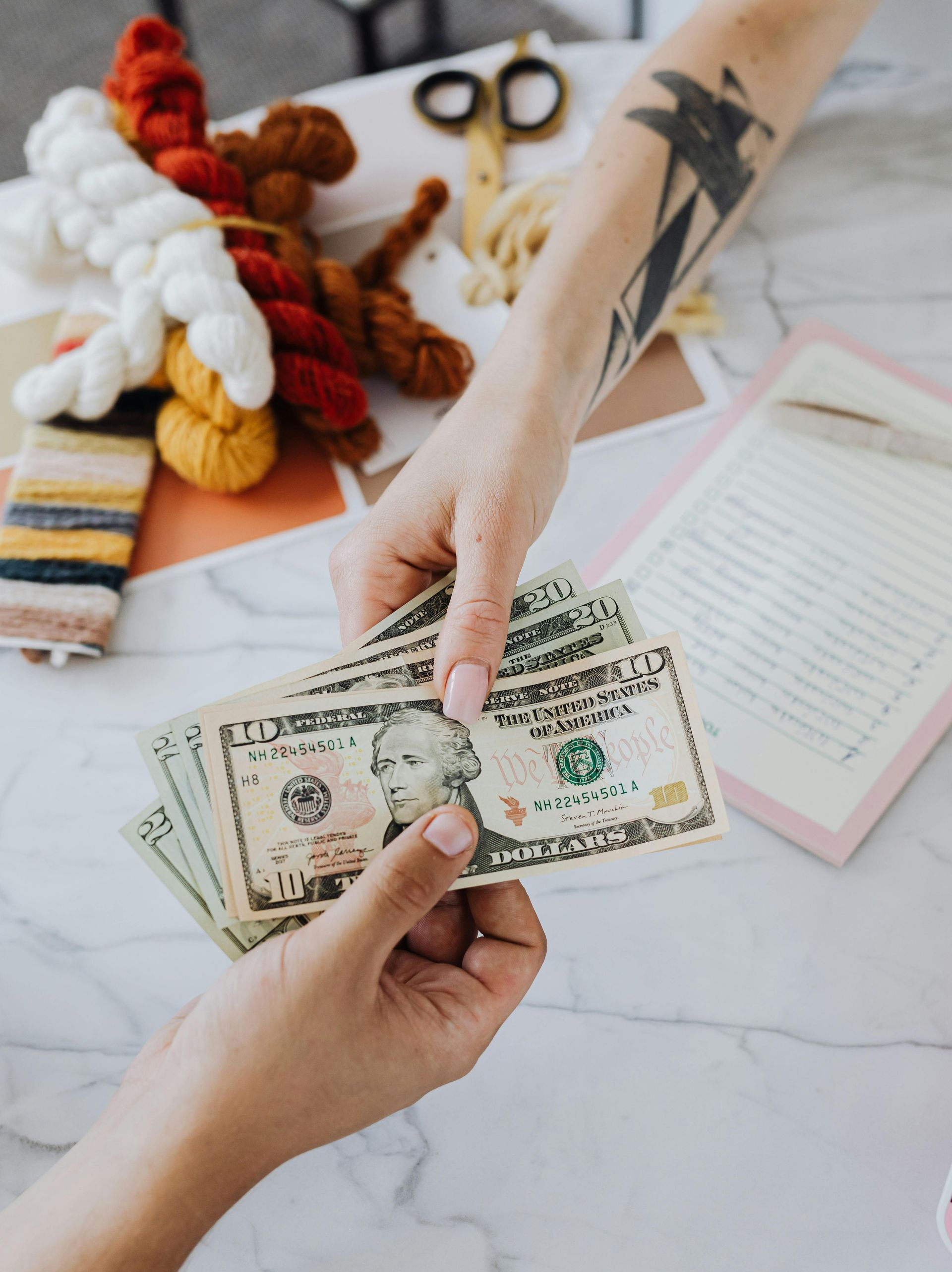 Hands exchanging US dollar bills over a white table with craft supplies.