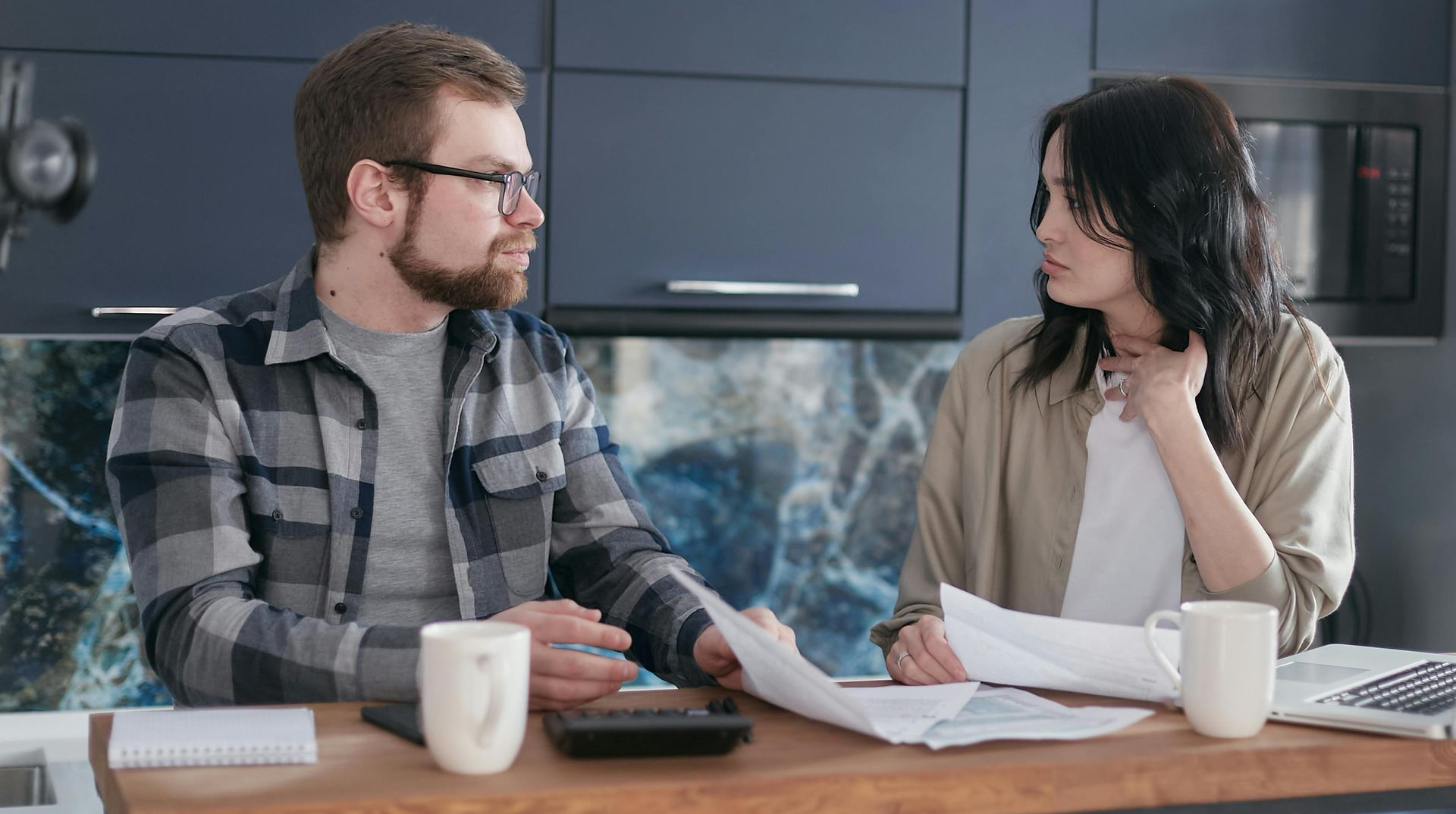 Man and woman reviewing bills in a kitchen, expressions appear concerned.