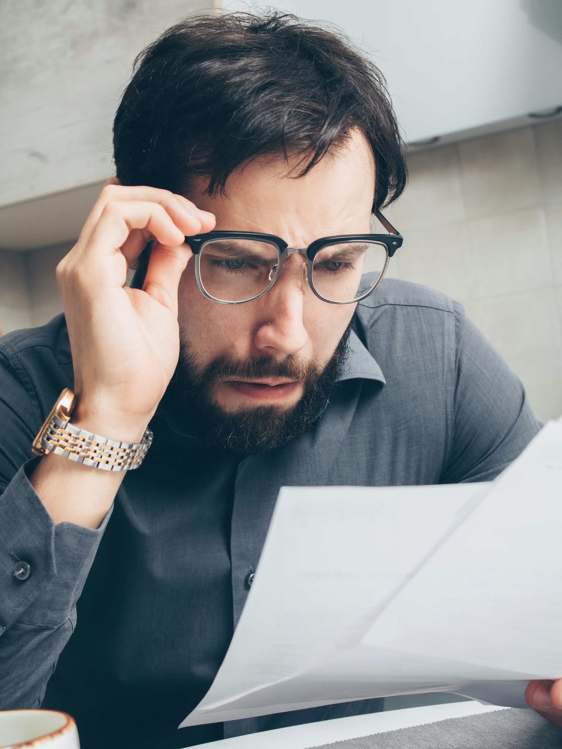 Man with glasses examines papers with a puzzled expression; holding glasses.