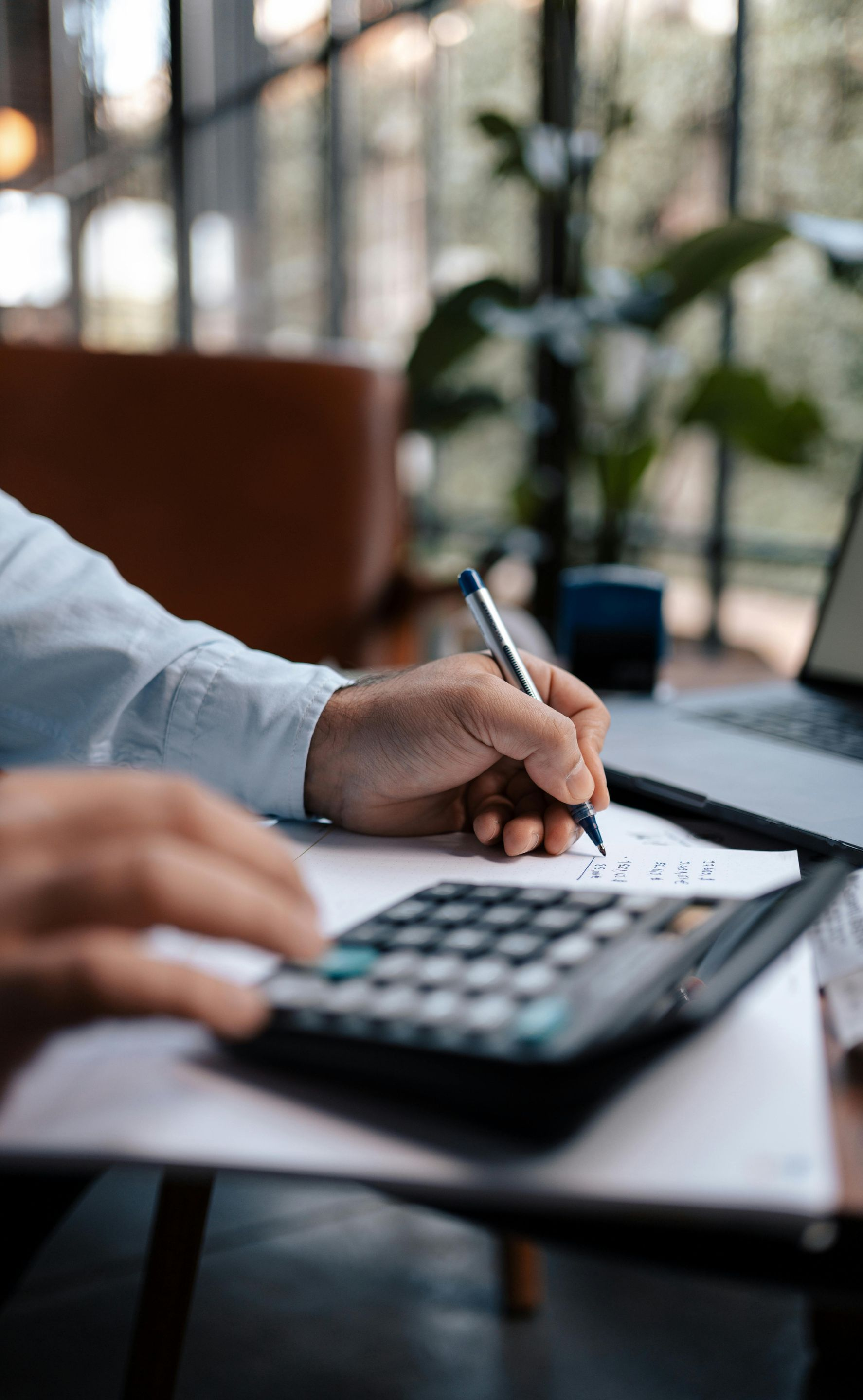 Person using a calculator and pen, working at a table with a laptop and documents, likely in an office setting.
