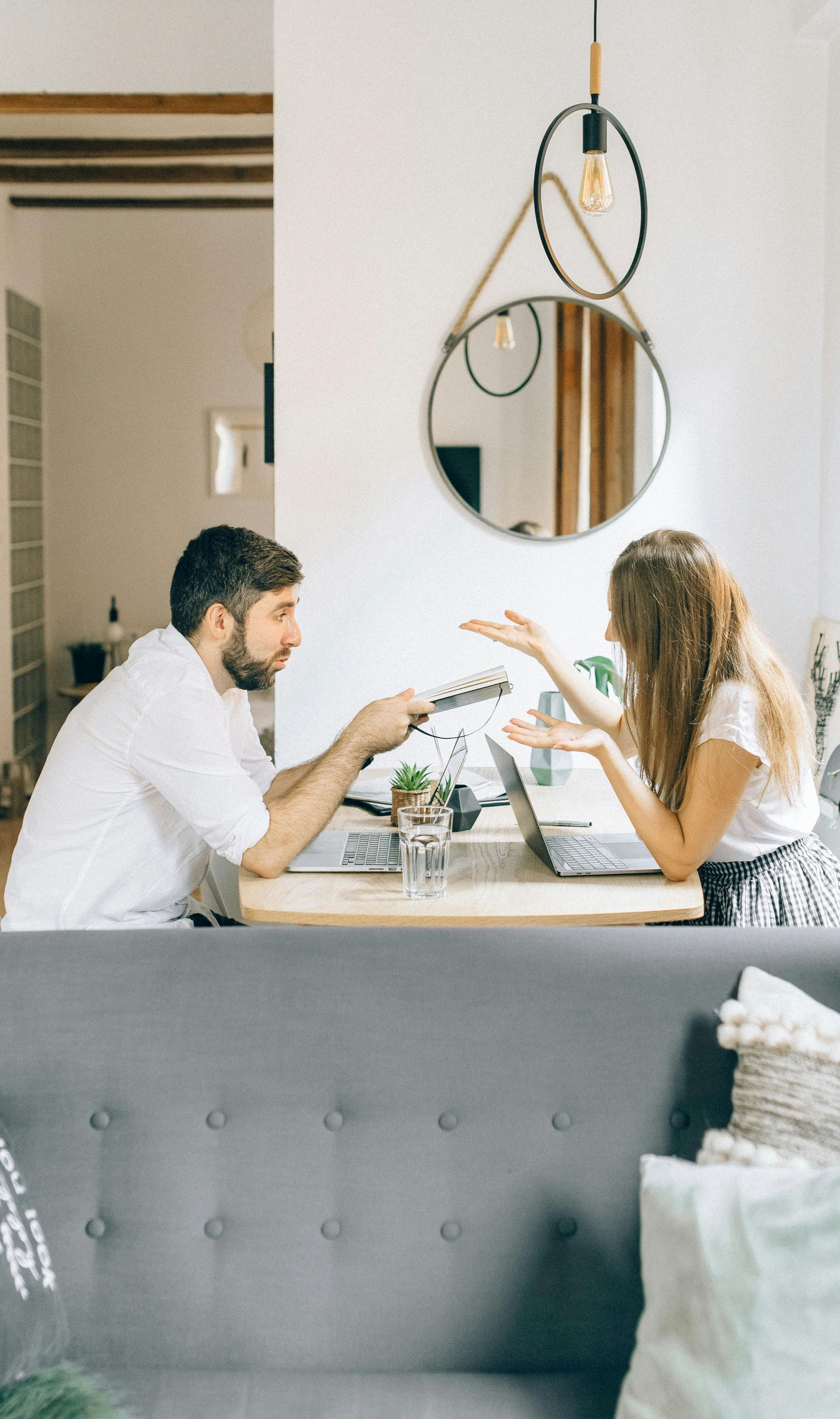 Man and woman working at a table with laptops, arguing, in a modern living room.