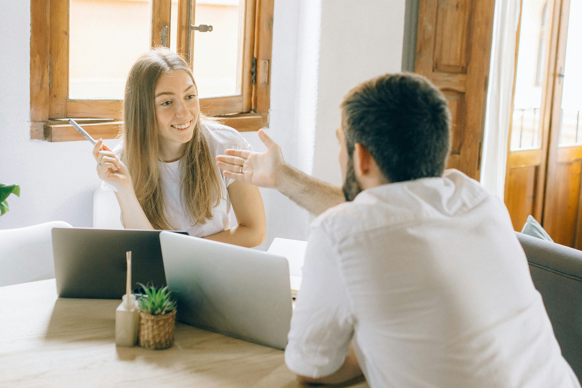 Woman and man at a table with laptops; woman smiles and gestures, man gestures back.