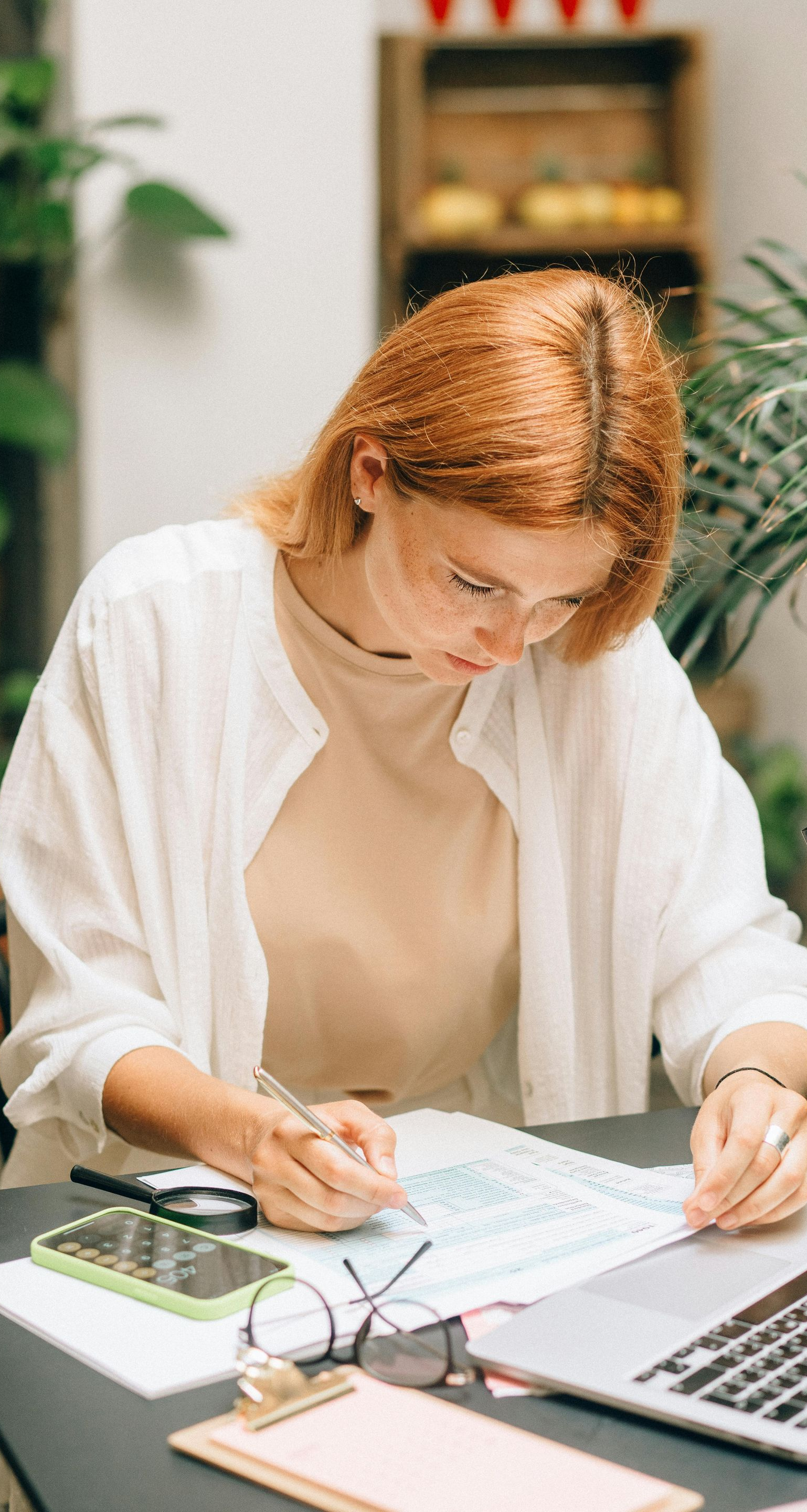 Woman with red hair writing at a table with laptop, phone, and glasses; plants in background.