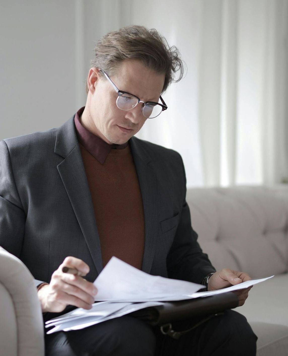 A man in a suit and glasses is sitting on a couch looking at papers
