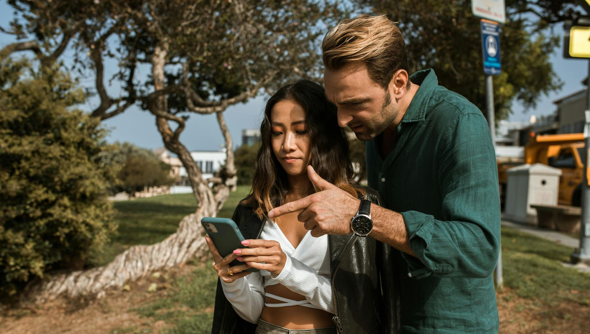 A woman and man looking at a phone outdoors. Man points to screen. Green trees and buildings in background.