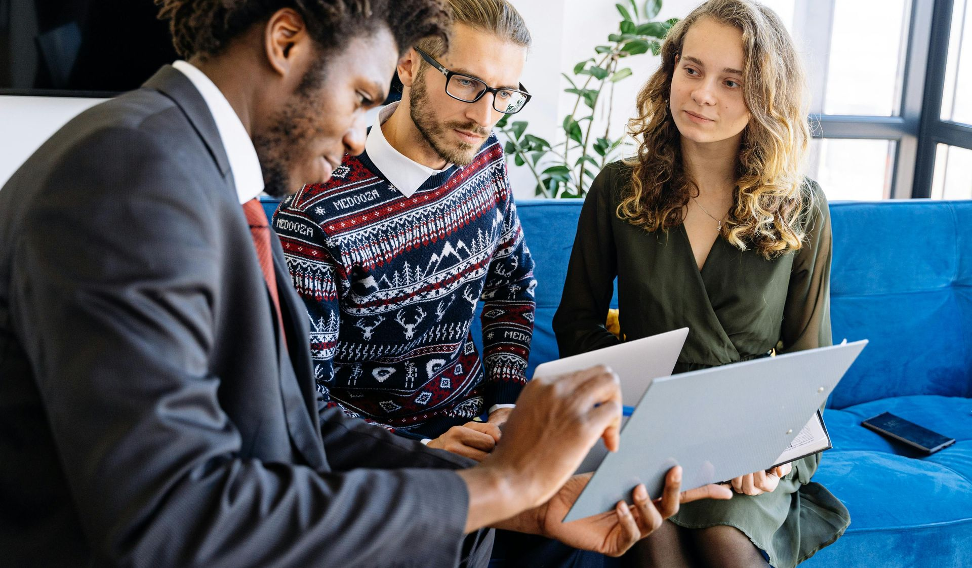Three people on a blue couch looking at documents. A man in a suit points.