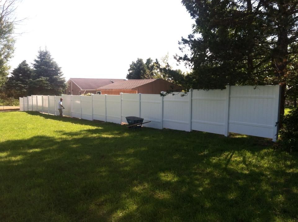 A white fence surrounds a lush green yard with a wheelbarrow in the foreground.