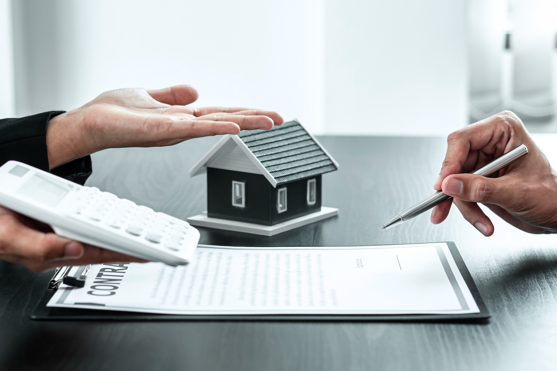 Hands around a model house and contract; one person signing, the other holding a calculator.