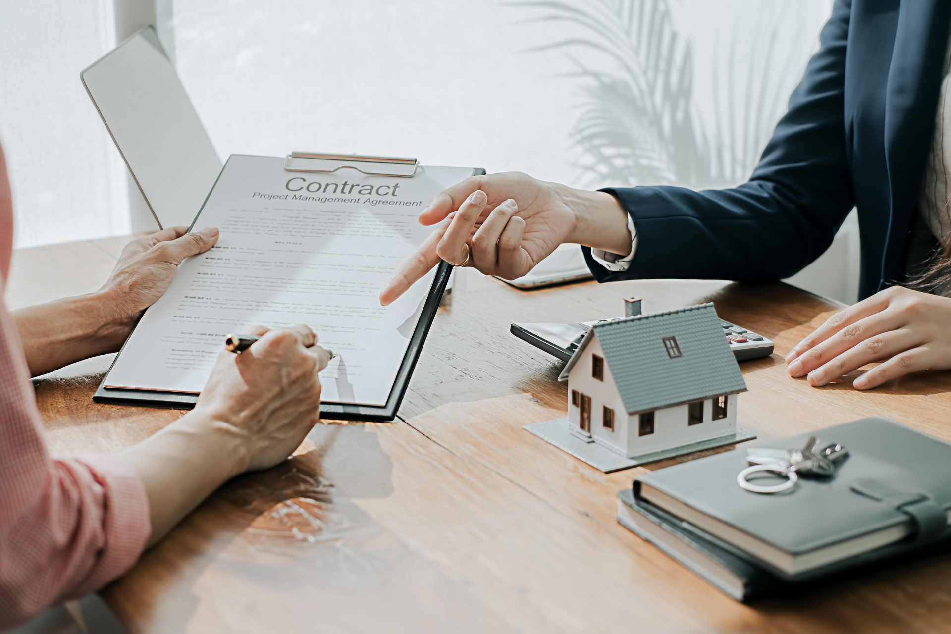Person signing a contract, guided by another. A small house model and keys on a desk.