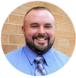Man with a shaved head smiles, wearing a blue shirt and patterned tie, in front of a brick wall.