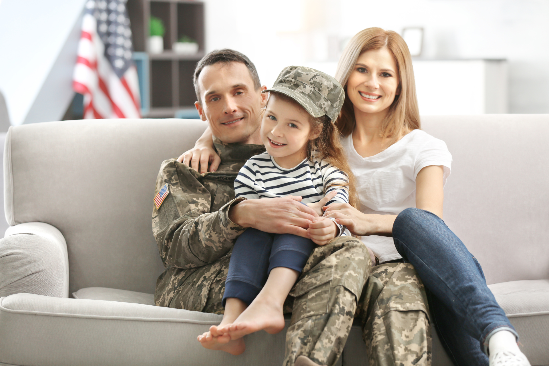 Family of three (soldier, woman, child) smiles on a couch, indoors. American flag in background.
