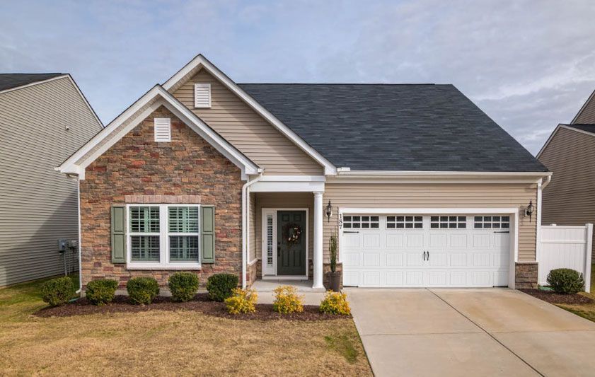 Bungalow-style house with stone and tan siding, white garage door, and a driveway on a sunny day.