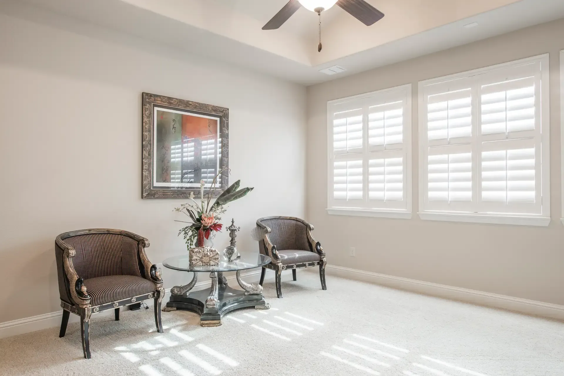 Two chairs, a glass table with decor, and a framed mirror in a beige-colored room with white window shutters.