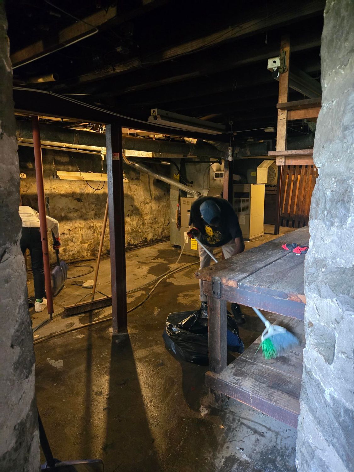 A person works in a dark, cluttered basement with stone walls and exposed beams. They are near a wooden structure and a trash bag.