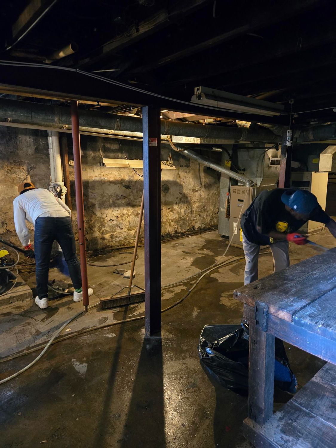 Two people working in a dark basement, likely on a renovation. One cuts a beam, while the other stands near a furnace.