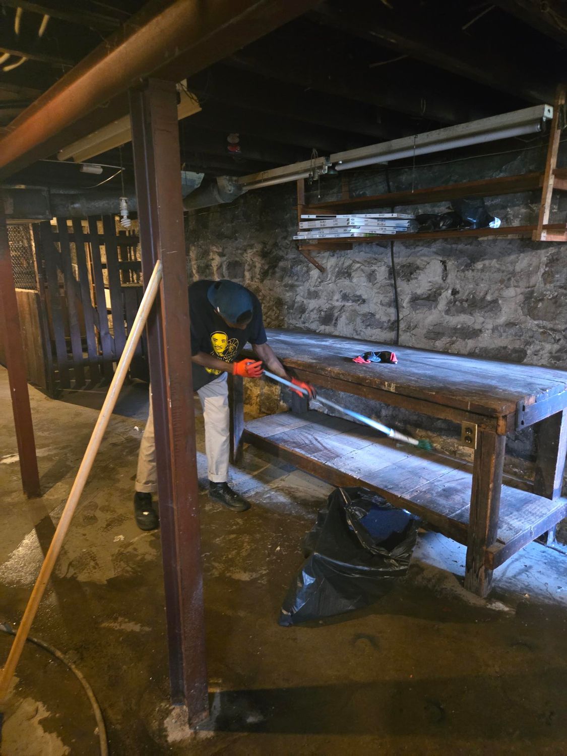 Person using a power tool on a workbench in a dimly lit basement. Stone wall and wooden structures visible.