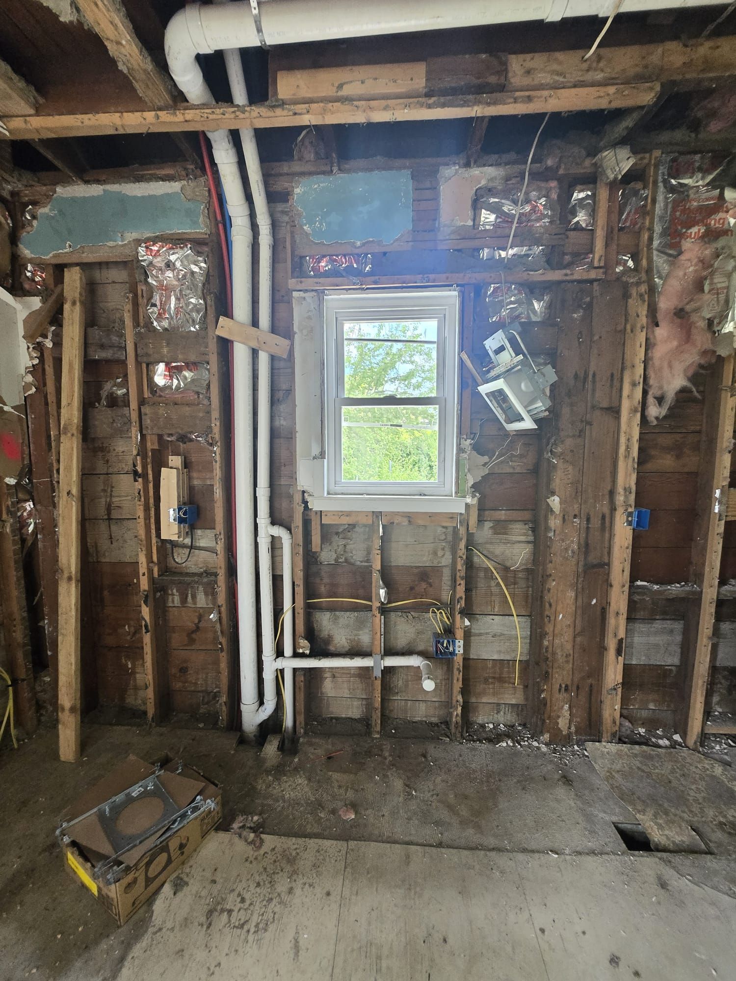 Interior wall of a home under renovation, exposing studs, pipes, and a window. The space is unfinished with visible electrical wiring and water lines.