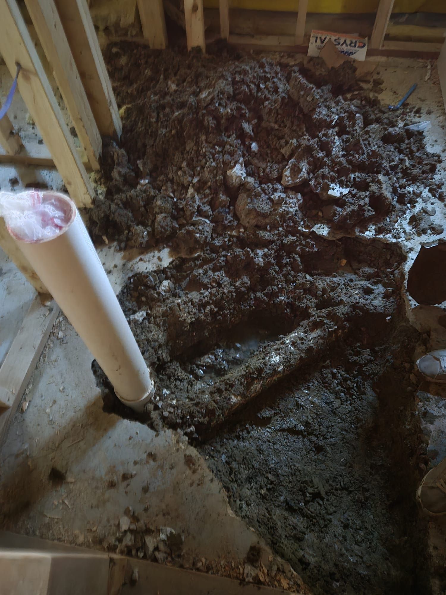 A plumbing job in progress. A white PVC pipe emerges from a concrete floor covered in dirt and debris.