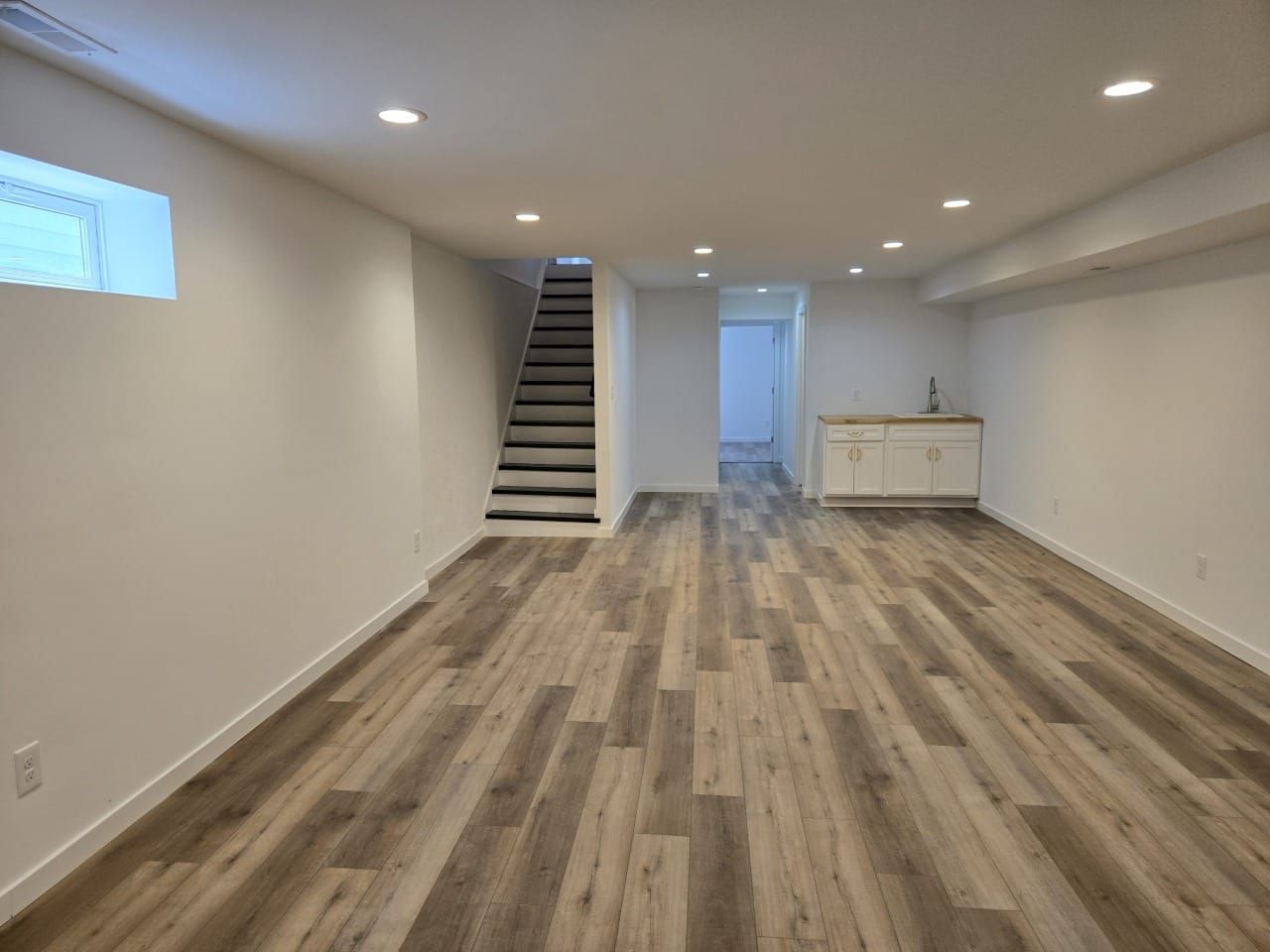 Empty finished basement with gray wood-look flooring, white walls, recessed lighting, stairs, and a small sink area.