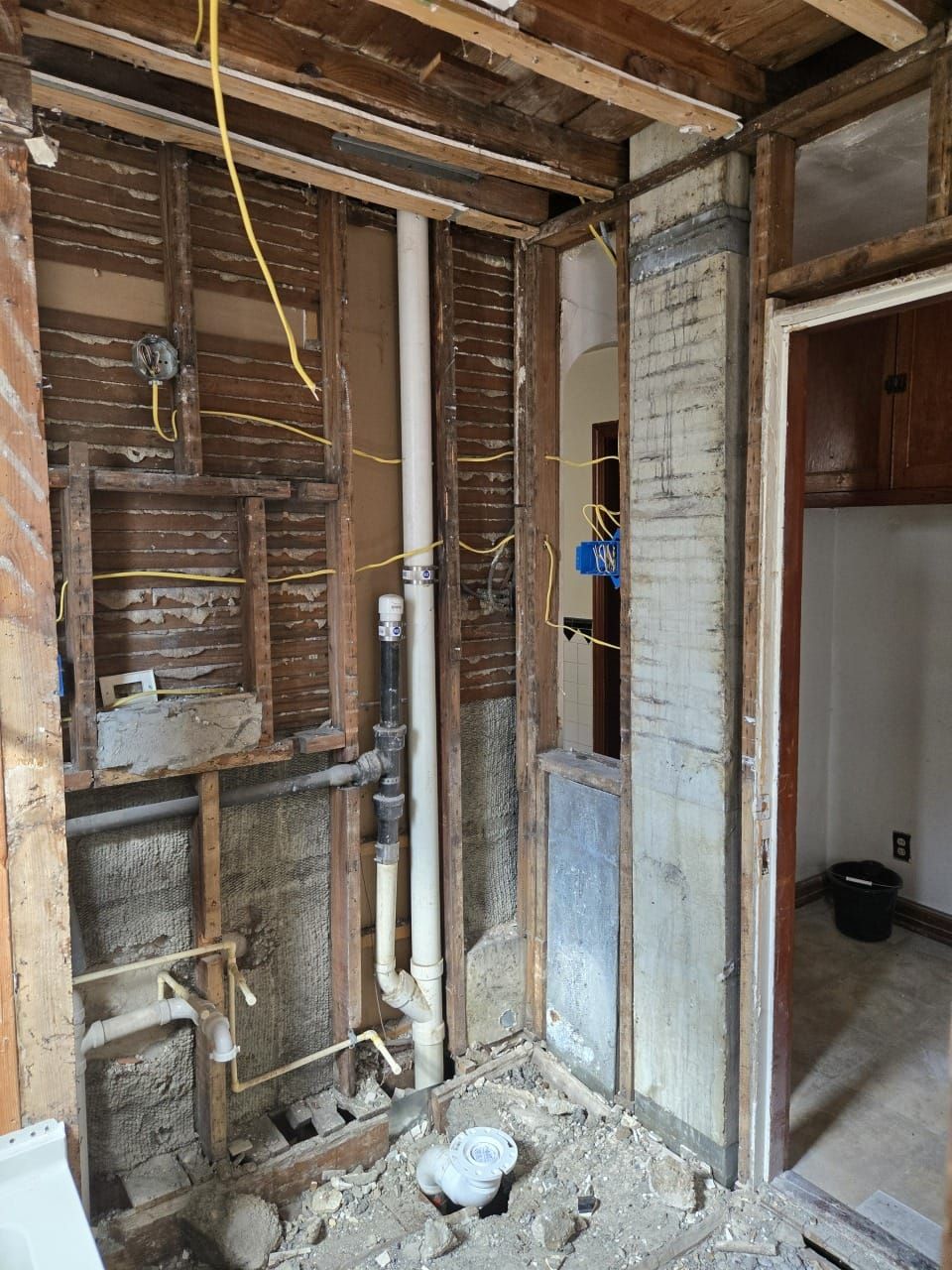 Interior view of a bathroom under renovation, exposing framing, pipes, and electrical wiring. A doorway leads to another room.