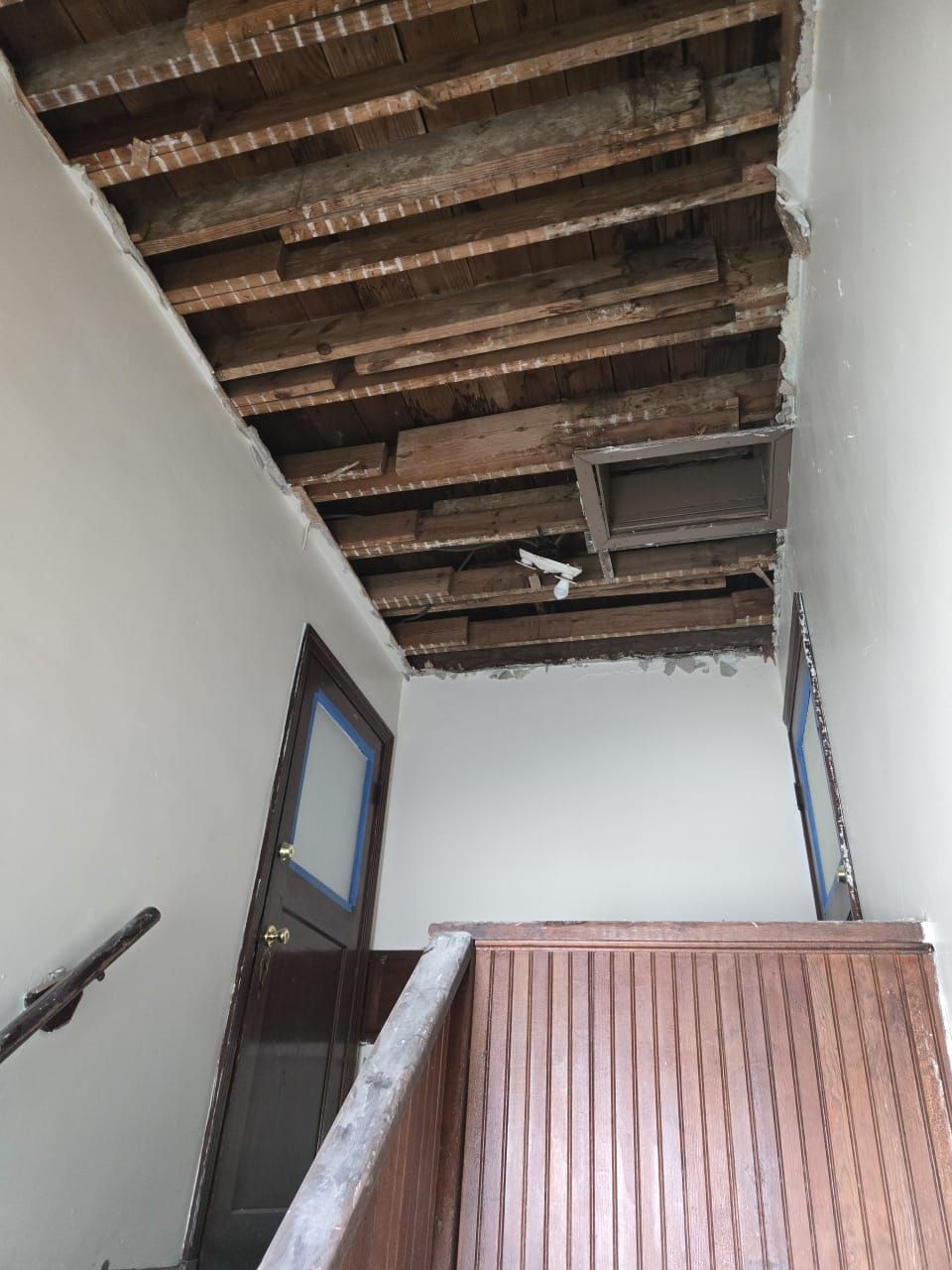 A view of a partially demolished ceiling above a staircase. Exposed wooden beams are visible, along with a door, and a partial handrail.