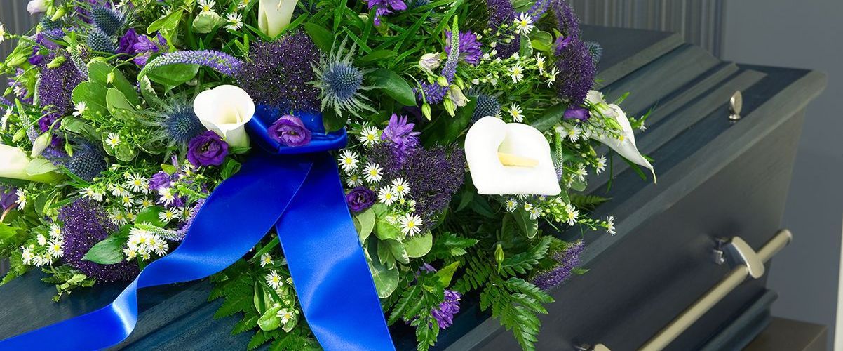 A coffin is decorated with purple and white flowers and a blue ribbon.