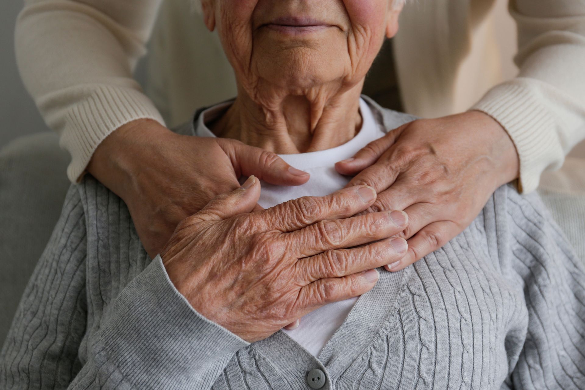 An elderly woman's hands held gently by another person's hands, both resting on her shoulders.