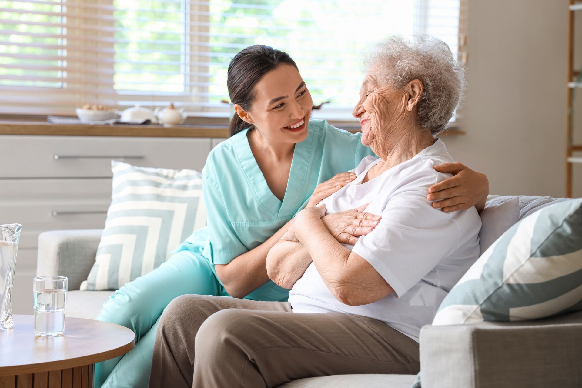 Caregiver in blue scrubs embraces elderly woman on a sofa indoors.