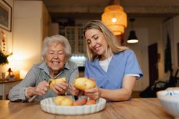 Woman and elderly person peeling potatoes together at a wooden table; both smiling.