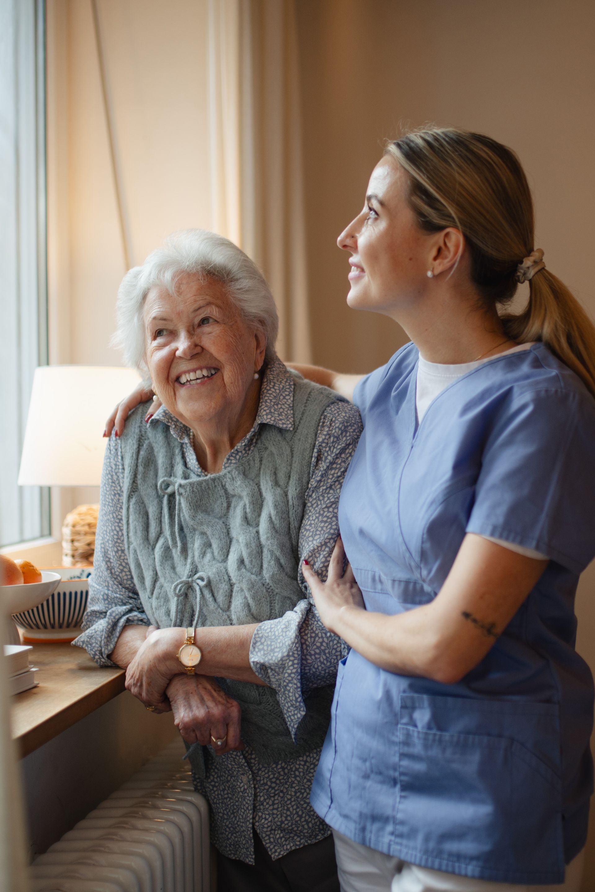 Woman in blue scrubs assists elderly woman looking out window, smiling.