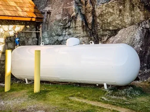 A large white propane tank is sitting in the grass in front of a rock wall.
